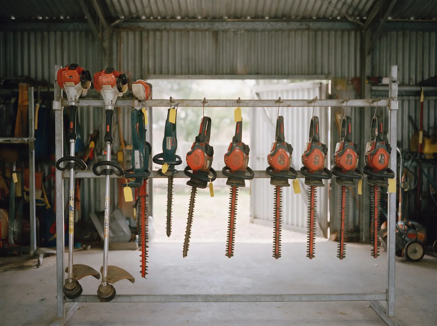 Rack of whipper snippers and hedge trimmers with yellow hire tags inside the shed