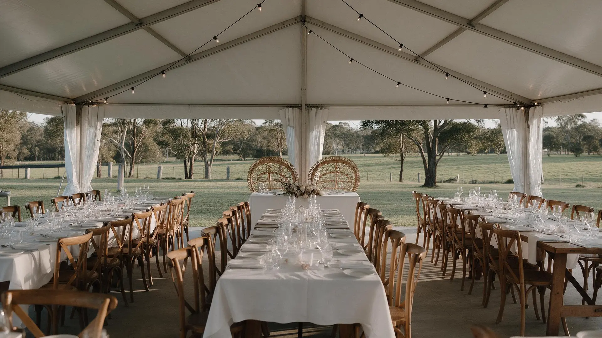 Long tables with white linen, chairs and a peacock-chair bridal table set under festoon lights inside an open-sided marquee