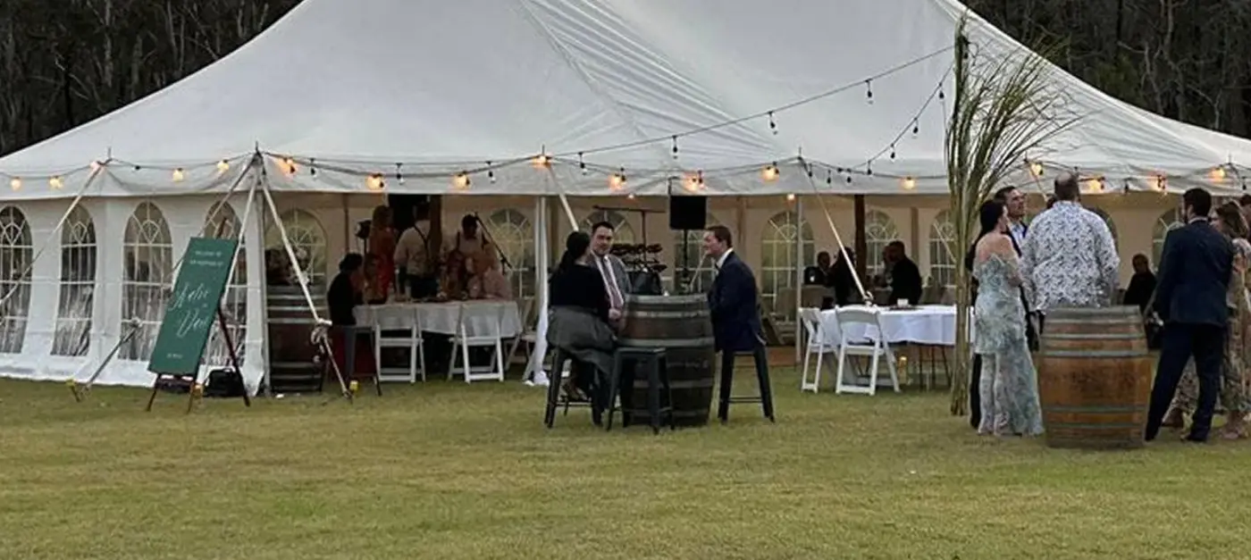 Guests gathered around wine barrels outside a peg-and-pole marquee at dusk with festoon lights and gum trees