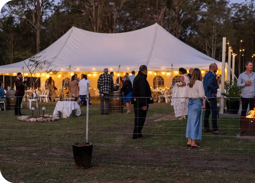 Wedding guests outside a glowing peg-and-pole marquee at dusk with gum trees behind