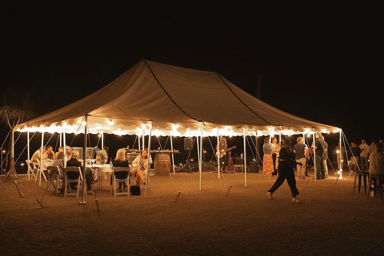 Grafton Hire peg-and-pole marquee at night with festoon lights, guests seated at tables and a musician playing
