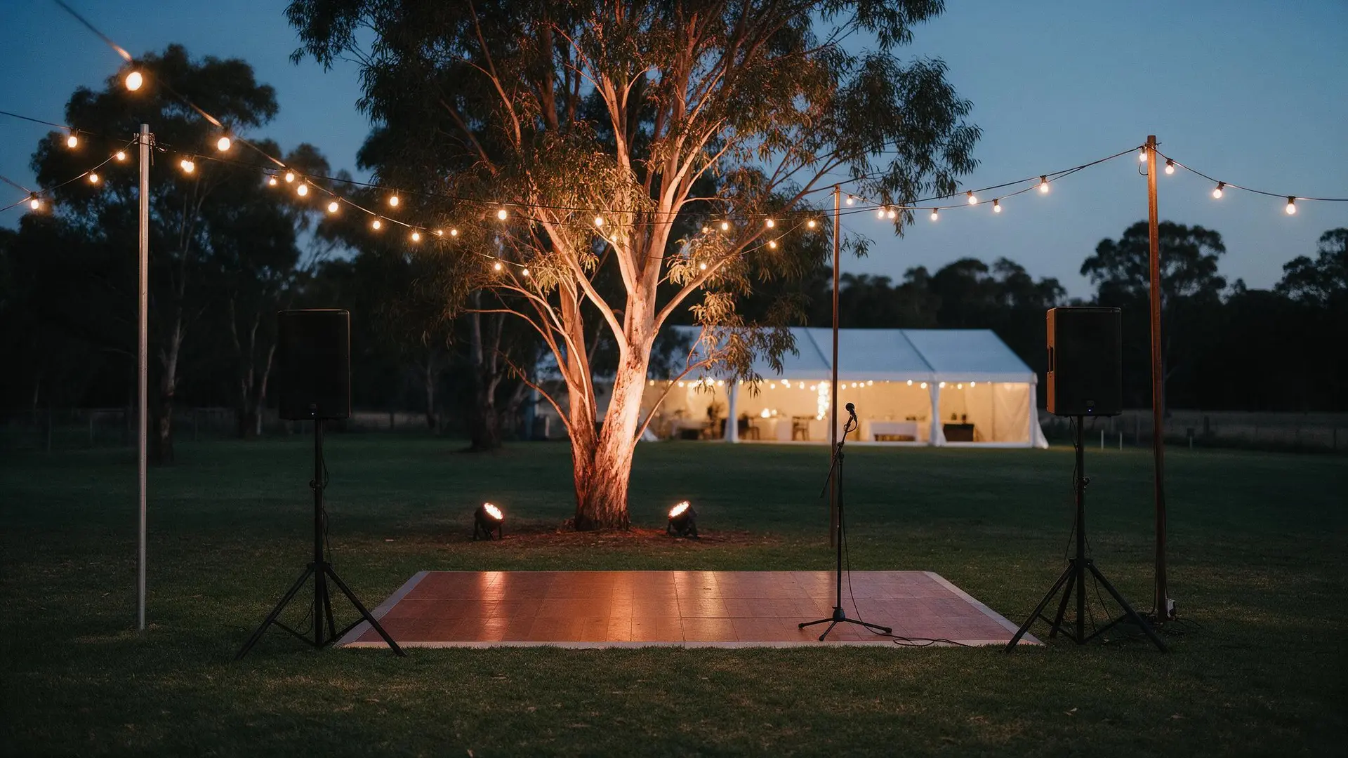 Festoon lights strung over a timber dance floor with PA speakers on stands, a lit gum tree and a white marquee behind at dusk