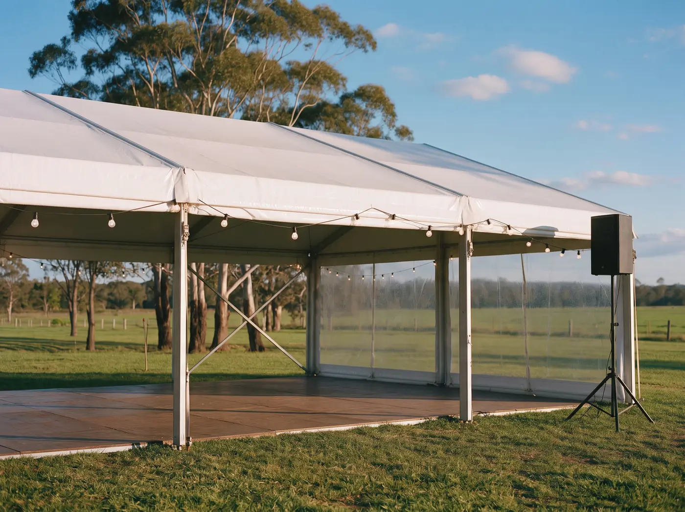 White marquee with festoon lights along the eave, a timber dance floor and a PA speaker on a tripod stand in a paddock at golden hour